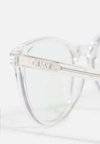 Lunettes de soleil en plastique transparent avec une monture ronde, présentant une branche argentée sur laquelle est gravé "QUAY" en lettres en relief.