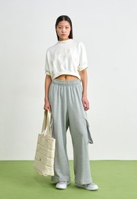 Sweatshirt cropped branco com o logótipo "Nike", calças de algodão cargo cinza, bolsa de lona acolchoada bege e ténis cinza-claro em piso verde.