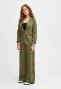 Olivově zelený blazer s přiléhavým střihem, spárovaný s kalhotami s širokými nohavicemi a bílými bočními pruhy. Hladká tkanina, neformální, ale elegantní vzhled.