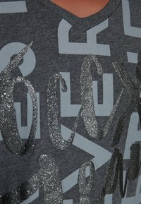 Šedé tričko s V výstřihem, které má velké, texturované stříbrné třpytivé písmena a grafické vzory ve světlejší šedé. Měkký bavlněný materiál.