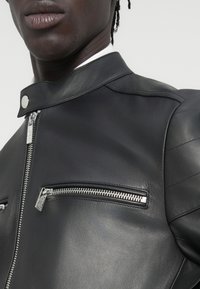 Μαύρο δερμάτινο μπουφάν με ψηλό γιακά, ασημένιο φερμουάρ και δύο τσέπες με φερμουάρ στο μπροστινό μέρος, με λεία υφή και κομψό σχεδιασμό.