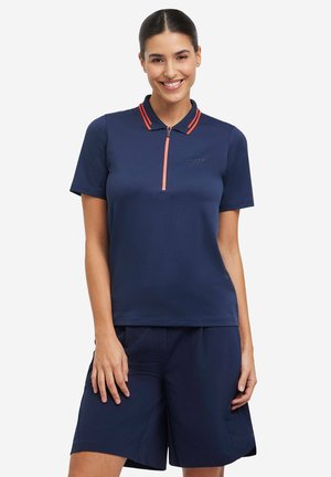 Kobieta uśmiechająca się, ubrana w granatową koszulkę polo z krótkim rękawem, czerwoną lamówką i przednim zamkiem błyskawicznym, w zestawie z dopasowanymi granatowymi szortami.