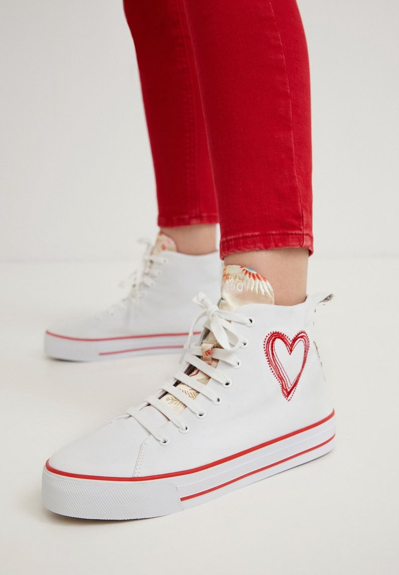 White high-top sneakers with red accents and a heart embroidery on the side. Textured tongue features a colorful pattern. Rubber sole.