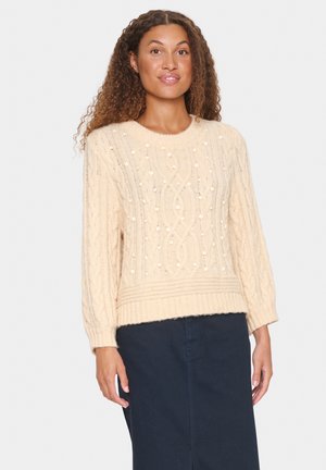 Krämfärgad kabelstickad tröja med texturerade mönster och bollar, rund hals och ribbad nederkant. Stylerad med en blå kjol.