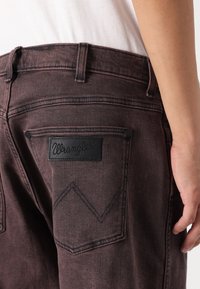 Tamnočvino crvene traperice s crnom kožnom oznakom Wrangler na stražnjem džepu, s ušivenim cik-cak uzorkom i bočnim šavovima.