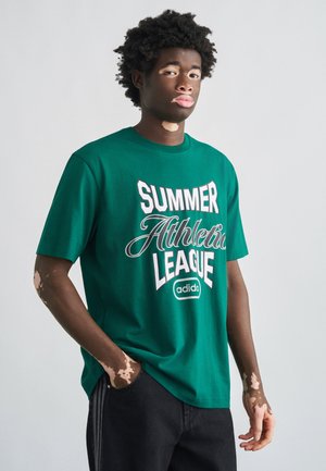 Zöld pamut rövid ujjú póló élénk fehér "SUMMER Athletic LEAGUE" felirattal és logóval. Fekete farmerrel viselve.