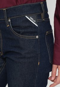 Tmavomodré rifle so slim fit strihom, kontrastným žltým prešívaním a bielou nášivkou "REPLAY" na prednom vrecku.