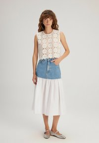 Un crop top en dentelle blanche avec des motifs circulaires, associé à une jupe en denim bleu et une partie inférieure plissée blanche. Des chaussures argentées complètent le look.