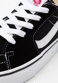 Baskets basses noires et blanches avec des lacets blancs, une bande latérale blanche et un logo rouge sur la languette, montrant des coutures détaillées et une semelle en caoutchouc.