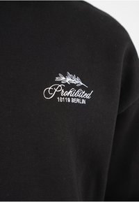 Sudadera de algodón negra con texto bordado en blanco "Prohibido" y "10119 BERLÍN", con un diseño de hoja como acento. Textura suave.