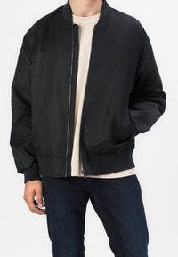 Svart bomberjacka med dragkedja fram, ribbade ärmslut och midja. Har en slät, glänsande yta. Bärs över en ljusbeige skjorta och mörka jeans.