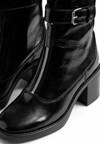 Botas de tobillo de cuero sintético negro con punta cuadrada, cremallera plateada y correa ajustable con hebilla. Presentan un tacón grueso y una textura suave.