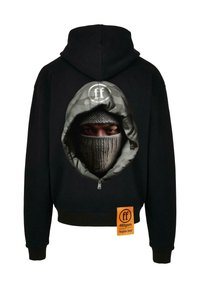 Sudadera negra con un gráfico de una capucha de camuflaje y una balaclava de punto que cubre el rostro de una persona, y una etiqueta naranja que dice "caras olvidadas" en el frente.