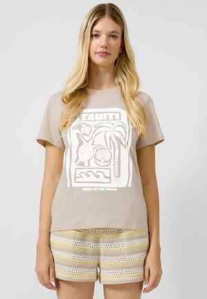 Młoda kobieta o długich blond włosach, ubrana w beżowy t-shirt z tropikalnym wzorem oraz pasiastymi dzianinowymi szortami, stojąca na jednolitym tle.