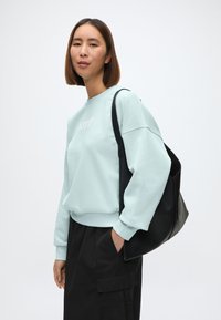 Gaiši zils sporta krekls ar ribotām piedurknēm, uz kura krūtīm ir neliels balts logo, kopā ar melniem biksēm un melnu somu.