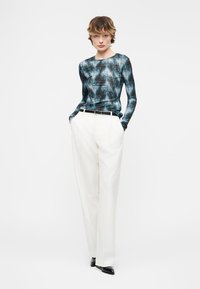 Άτομο που στέκεται φορώντας μπλε μακρυμάνικη μπλούζα με σχέδια, φαρδιά λευκά παντελόνια ψηλόμεσα, μαύρη ζώνη και μαύρα παπούτσια.