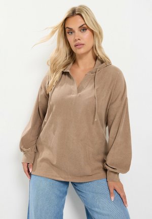 Femme blonde portant un sweat à capuche ample beige avec des cordons et un jean bleu clair, debout devant un fond blanc uni.