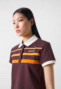 Bordo polo majica s bijelim ovratnikom, s horizontalnim narančastim prugama i izvezenim bijelim logom na prsima. Kratki rukavi.