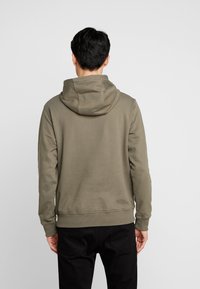 Uomo di spalle che indossa un semplice hoodie verde oliva e pantaloni neri su uno sfondo grigio neutro.