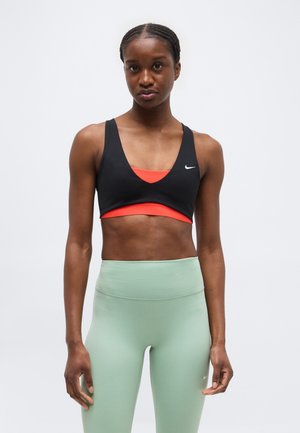 Atletisk kvinde med flettet hår iført sort og orange sports-bh og lysegrønne, højtaljede leggings, stående mod en ensfarvet baggrund.