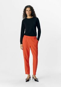 Haut noir à manches longues, pantalon rouge ajusté avec poches et escarpins noirs à bout pointu. Tissu lisse avec une silhouette ajustée.