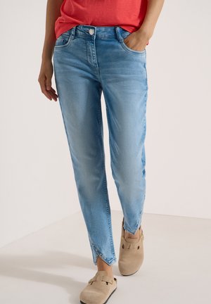 Femme portant un jean bleu clair avec des ourlets croisés et des chaussures beige à enfiler, debout avec une main dans la poche et un haut rouge partiellement visible.