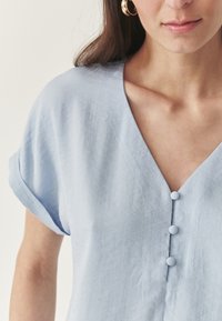 Blusa di lino di colore azzurro chiaro con scollatura a V, maniche corte arrotolate e tre bottoni rivestiti in tessuto dello stesso colore lungo la parte anteriore.