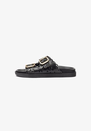 Black sandals with two adjustable buckles, floral cut-out design, flat sole, and smooth texture. Minimalist and functional aesthetic.