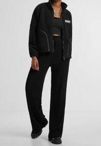 Chaqueta de forro polar negra con cremallera, con ribetes blancos y bolsillos laterales, combinada con pantalones anchos de pierna acanalados negros a juego y zapatos negros gruesos.