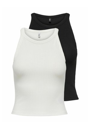 Ung kvinne står med hånden i lommen på mørke vide jeans, iført en hvit ermeløs topp og svarte åpne hæler på ensfarget bakgrunn.
