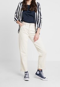 Pruhovaný námořnický a bílý blazer přes námořnické tričko, spárovaný s bílými kalhotami s širokými nohavicemi a modrými vysokými teniskami s bílou podešví.