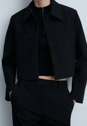 Personne portant une veste noire courte avec de grandes poches avant sur un haut noir côtelé à fermeture éclair et un pantalon noir assorti, les mains dans les poches.