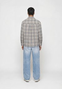 Άτομο που φοράει μπεζ καρό πουκάμισο με μακριά μανίκια, φωτεινό μπλε φαρδιά τζιν και λευκά αθλητικά παπούτσια, στέκεται με πλάτη προς το μέρος μας μπροστά σε μια απλή φόντο.