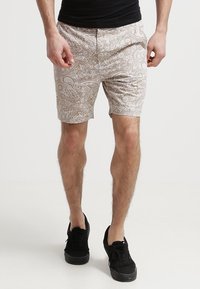 Shorts beiges à motifs avec des designs complexes, présentant un devant plat et des poches, associés à des chaussures noires.