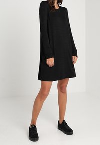Abito nero a maniche lunghe con vestibilità rilassata, scollatura rotonda e tessuto morbido, abbinato a sneakers nere. Design minimalista, senza motivi.