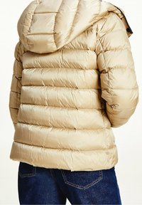 Beige pufferjacka med huva, med ett quiltat mönster och slät textur. Bärs över mörkblå jeans.