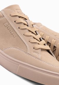 Béžové semišové tenisky s plochými tkaničkami, kontrastním prošíváním, texturovanou gumovou podrážkou a vyraženým logem na boku.