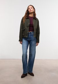 Donna in piedi con capelli lunghi e castani, indossa una giacca verde oliva, un top bordeaux, jeans blu e stivali di camoscio marroni su uno sfondo neutro.