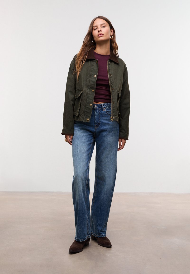 Donna in piedi con capelli lunghi e castani, indossa una giacca verde oliva, un top bordeaux, jeans blu e stivali di camoscio marroni su uno sfondo neutro.