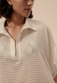 Lekka, przezroczysta bluzka w off-white z poziomymi paskami, klasycznym kołnierzykiem i zapięciem na przodzie. Miękka faktura i luźny krój.
