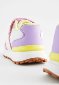 Baskets sportives présentant un corps en tissu blanc avec des accents violets, roses et jaunes. La semelle est texturée avec une adhérence en caoutchouc marron.