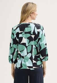 Blusa de corte holgado sobre un fondo negro, con un patrón geométrico en verde y blanco. Manga francesa con puños elásticos.