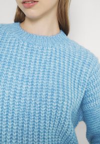 Světle modrý pletený svetr s žebrovanou strukturou, kulatým výstřihem a měkkým, prodyšným materiálem. Detailní záběr zdůrazňuje detaily šití.