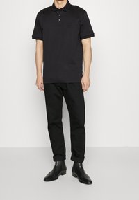 Svart pikétröja med korta ärmar, tre-knäpps knäppning och en liten bröstficka; kombinerad med svarta uppvikta byxor och ankelboots.