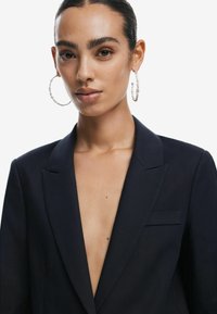 Sötétkék blézer mély V-kivágással, sima anyagból készült. A modellen nagy, texturált karika fülbevalók vannak; az apró zsebdíszítés a megjelenést finomítja.