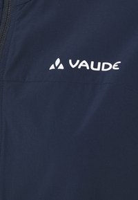 Veste Vaude bleu marine avec une texture lisse, une poche zippée et un logo blanc visible à l'avant. Légère, conçue pour une utilisation en extérieur.