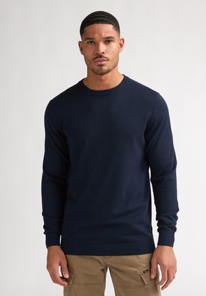 Mann trägt einen marineblauen Langarmpullover und beige Cargohosen, steht vor einem schlichten hellen Hintergrund.