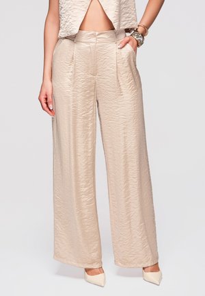 Kvinde iført beige højttaljede bukser med vidde og tekstur, med venstre hånd i lommen og beige spidse hæle.