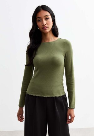 Top de manga larga de canalé en verde oliva con cuello redondo, dobladillo sin rematar y silueta ajustada, combinado con pantalones negros holgados.