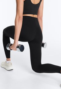 Kobieta w czarnym stroju sportowym wykonująca wykrok z chromowanymi hantlami, ubrana w białe trampki na białym tle.